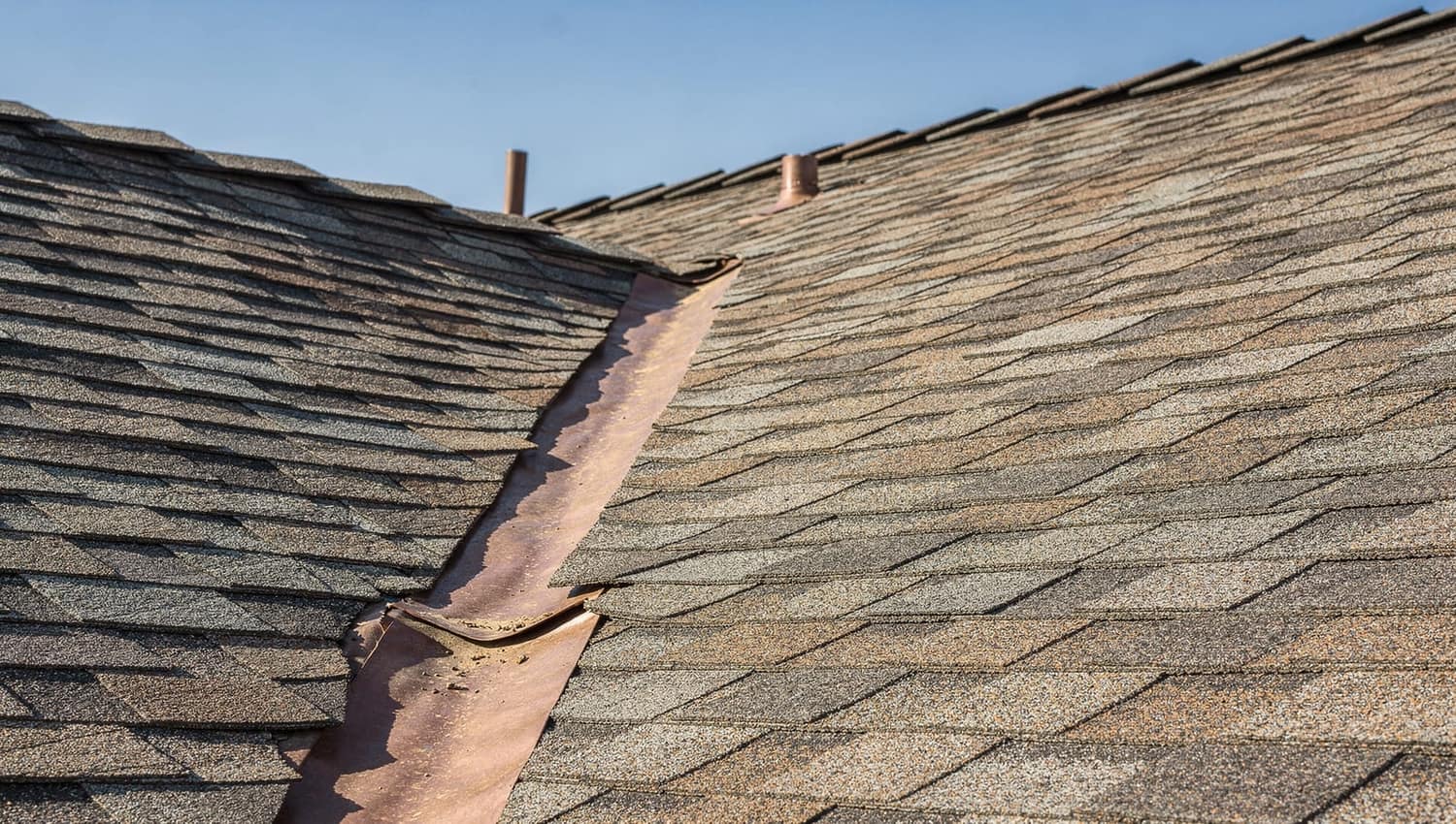 A close look at curling asphalt shingles and worn copper flashing along a roof valley